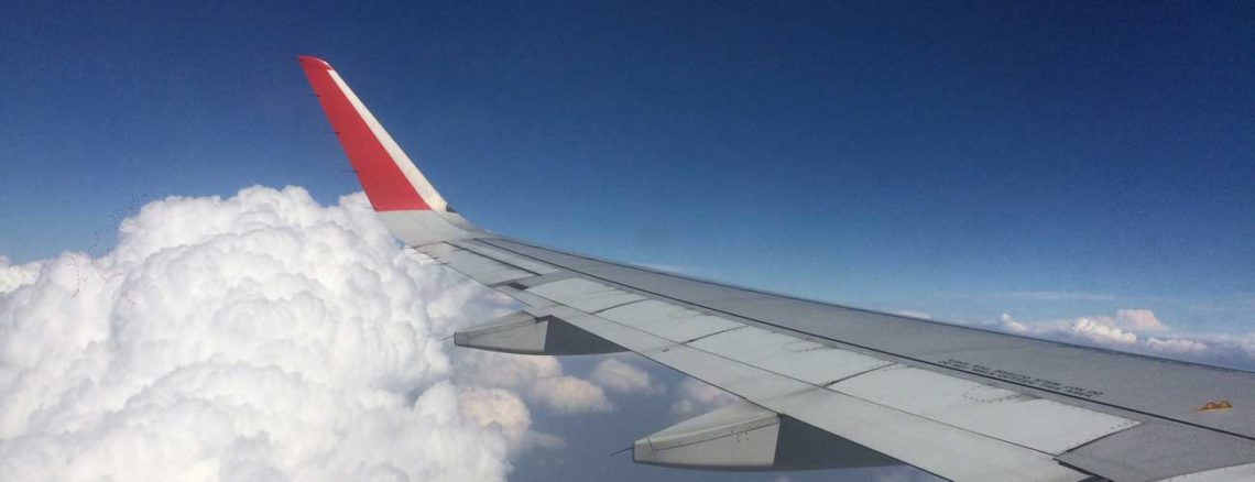 Image contains a photo of the wing of a plane with a blue sky above it and clouds below.