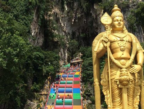 Image contains a photo of a large set of stairs ascending into a cave. The stairs are painted in bright colours and they're surrounded by jungle vegetation. In the foreground, to the right of the stirs, there is a large golden statue.