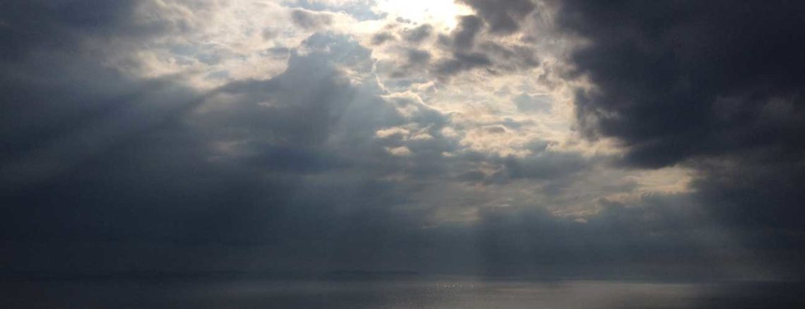 Image contains a photo of a large body of salt water extending beyond the horizon. Above the horizon line, there are clouds and a variety of sun beams breaking through them down to the water below.