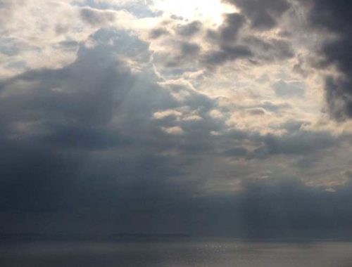 Image contains a photo of a large body of salt water extending beyond the horizon. Above the horizon line, there are clouds and a variety of sun beams breaking through them down to the water below.