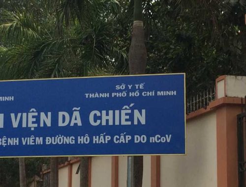 Image contains a photo of a large blue sign with white Vietnamese text. The sign is located just outside of a large wall and there are trees in the background.