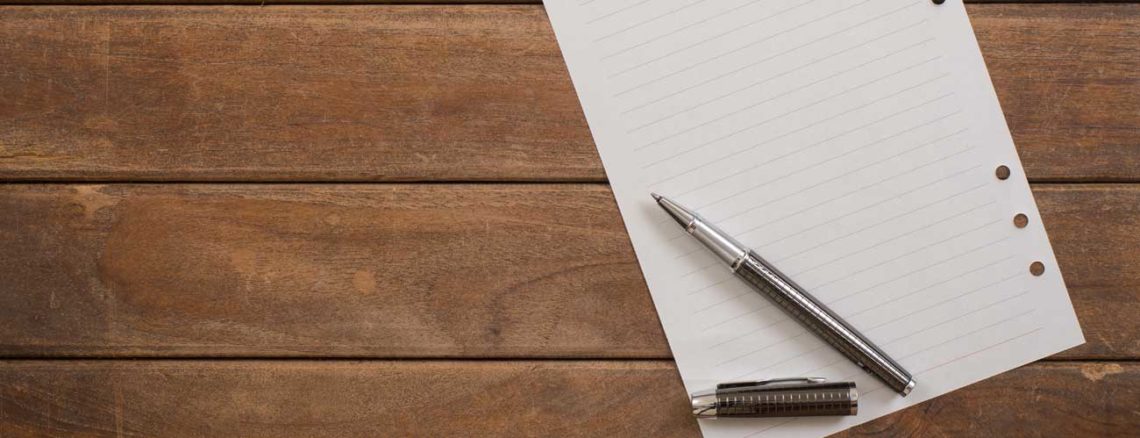 Image contains a photo of a piece of lined paper with holes punched on its right-hand side. A pen is placed at the bottom left-hand side of the paper, and its cap is resting just below it. The paper itself is placed on top of a wooden background consisting of three boards.
