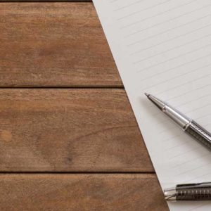 Image contains a photo of a piece of lined paper with holes punched on its right-hand side. A pen is placed at the bottom left-hand side of the paper, and its cap is resting just below it. The paper itself is placed on top of a wooden background consisting of three boards.