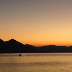 Image contains a photo of a sunset over a large body of water. On the horizon, hills can be seen stretching across most of the frame. The sky is a bright orange near the horizon, and it eventually turns a dark blue near the top of the frame.