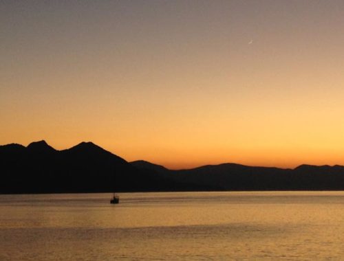 Image contains a photo of a sunset over a large body of water. On the horizon, hills can be seen stretching across most of the frame. The sky is a bright orange near the horizon, and it eventually turns a dark blue near the top of the frame.