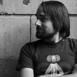 Image contains a black and white photo of a male looking sideways to the left of the camera. His arms are crossed and he's wearing a t-shirt. In the background, there is a large brick wall and a telephone pole to the right of the man. There are various posters on the pole.