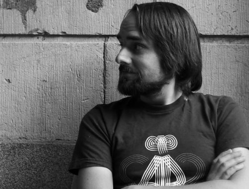 Image contains a black and white photo of a male looking sideways to the left of the camera. His arms are crossed and he's wearing a t-shirt. In the background, there is a large brick wall and a telephone pole to the right of the man. There are various posters on the pole.