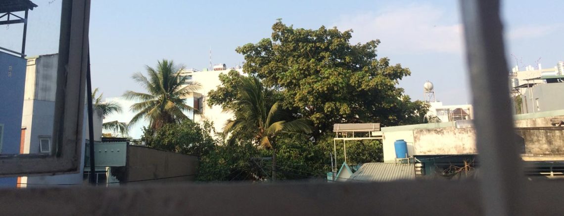 Image contains a photo from a bedroom window. White metal bars are visible in the foreground, and there is a blue sky and green trees in the background. Along with the trees, there are a number of small concrete houses.