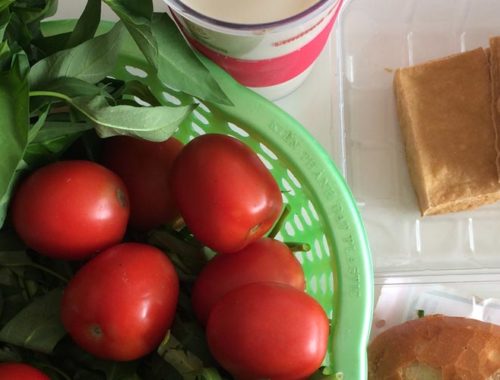 Image contains a photo of a variety of food on a white tabletop. On the left-hand side, in a green plastic basket, there are bright red tomatoes and a bunch of morning glory. To the right of the basket, there is a plastic container of small blocks of fried tofu, and just below that there is a banh mi sandwich.