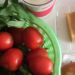 Image contains a photo of a variety of food on a white tabletop. On the left-hand side, in a green plastic basket, there are bright red tomatoes and a bunch of morning glory. To the right of the basket, there is a plastic container of small blocks of fried tofu, and just below that there is a banh mi sandwich.