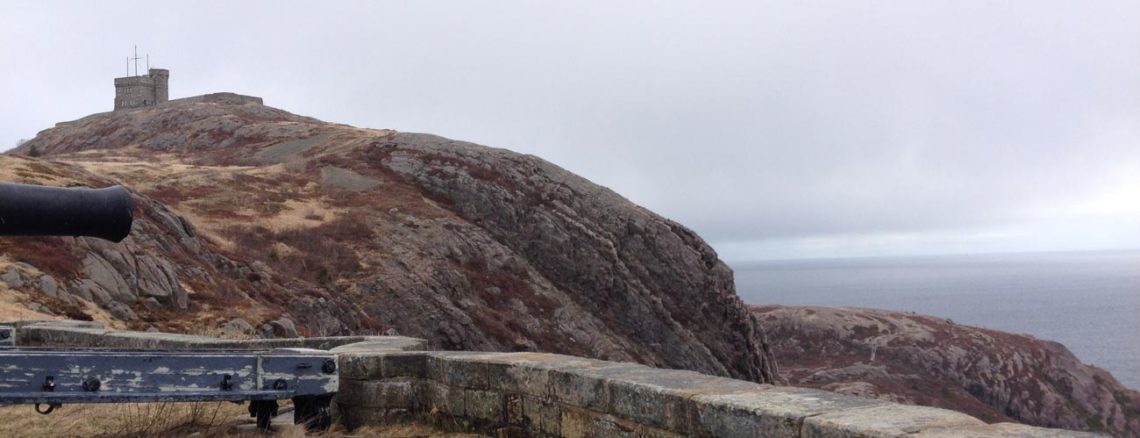 Image contains a photo of Signal Hill in St. John’s, Newfoundland and Labrador. In the foreground, an old cannon can be seen sticking out from the left, and there is a shot brick wall just in front of it. In the background, there is a hill with a small tower on top of it. Beyond that, there is a grey sky and the ocean.