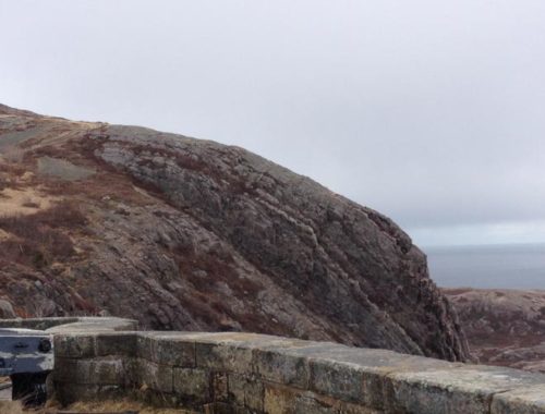 Image contains a photo of Signal Hill in St. John’s, Newfoundland and Labrador. In the foreground, an old cannon can be seen sticking out from the left, and there is a shot brick wall just in front of it. In the background, there is a hill with a small tower on top of it. Beyond that, there is a grey sky and the ocean.