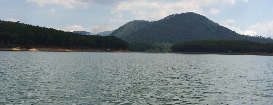 Image contains a photo of a relatively calm lake. In the background, there are rolling hills covered in tall green trees against a blue sky.