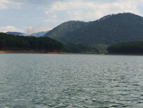 Image contains a photo of a relatively calm lake. In the background, there are rolling hills covered in tall green trees against a blue sky.