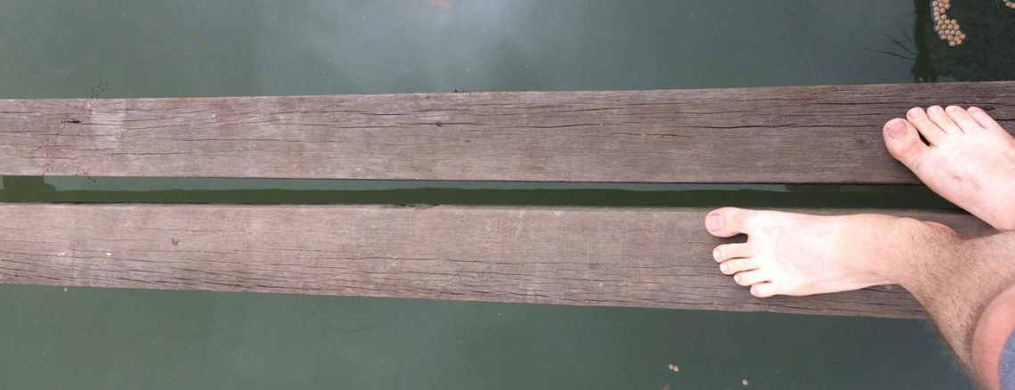 Image contains a photo of two thin wooden boards spanning the length of the image horizontally. Below the boards, there is dark green water. On the right-hand side of the image, two feet can be seen pointing across the boards.