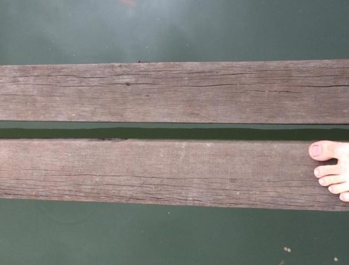 Image contains a photo of two thin wooden boards spanning the length of the image horizontally. Below the boards, there is dark green water. On the right-hand side of the image, two feet can be seen pointing across the boards.