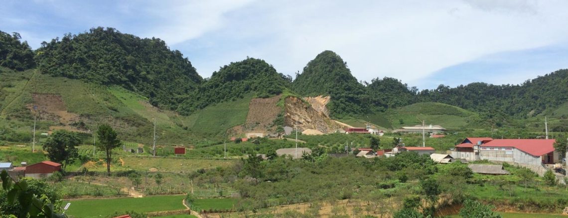 Image contains a photo of rolling hills and a green valley. In the foreground the peaks of several buildings with red tiled roofs can be seen.