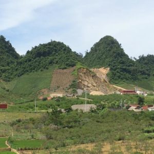 Image contains a photo of rolling hills and a green valley. In the foreground the peaks of several buildings with red tiled roofs can be seen.