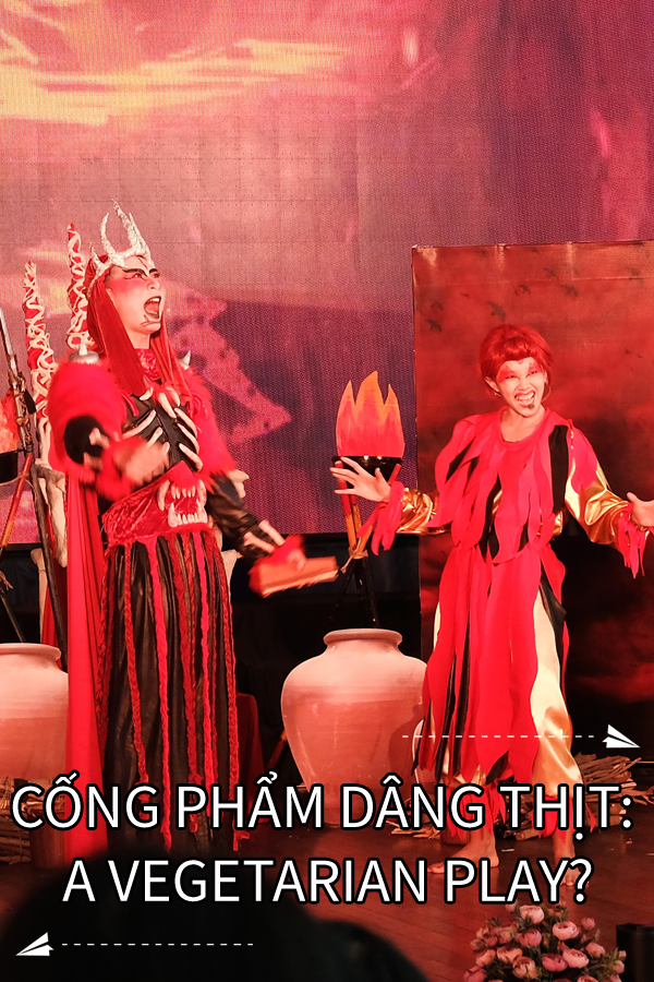Image contains a photo of a theatrical production involving two characters in a setting similar to Hell. the male character is taller and wears a crown and armour, as well as a cape. The female character is striking a comedic pose with a smile on her face. Much of the photo is in red tones. At the bottom of the image, there is white text in the centre that says "Cống Phẩm Dâng Thịt: A Vegetarian Play?". On the right-hand side above the text, there is a small white paper airplane with a dotted line behind it. Below the text and on the left-hand side, there is a similar icon.