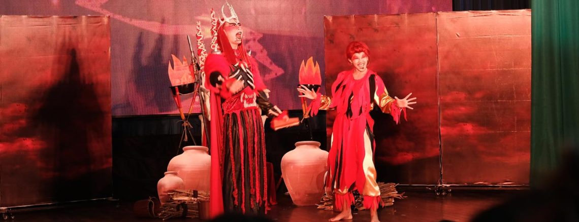 Image contains a photo of a theatrical production involving two characters in a setting similar to Hell. the male character is taller and wears a crown and armour, as well as a cape. The female character is striking a comedic pose with a smile on her face. Much of the photo is in red tones.