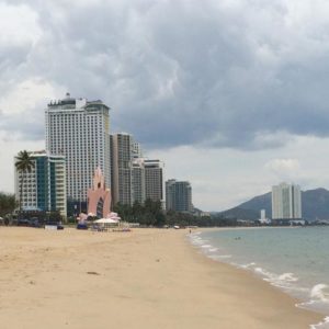 Image contains a photo of a beach with sand on the left and the ocean on the right. In the background, a number of large hotels can be seen.