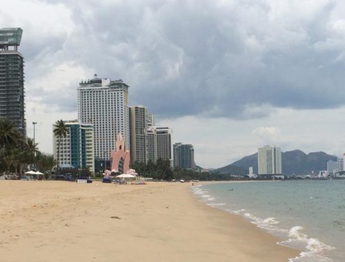 Image contains a photo of a beach with sand on the left and the ocean on the right. In the background, a number of large hotels can be seen.