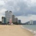 Image contains a photo of a beach with sand on the left and the ocean on the right. In the background, a number of large hotels can be seen.