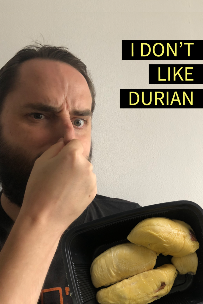 Image contains a photo of a bearded white man pinching his nose and looking unhappy. To the right of the man, there is a black plastic tray of durian, which is a yellowish fruit shaped in large pieces. In the top right-hand side of the image, there is yellow text on black strips that says "I Don't Like Durian".