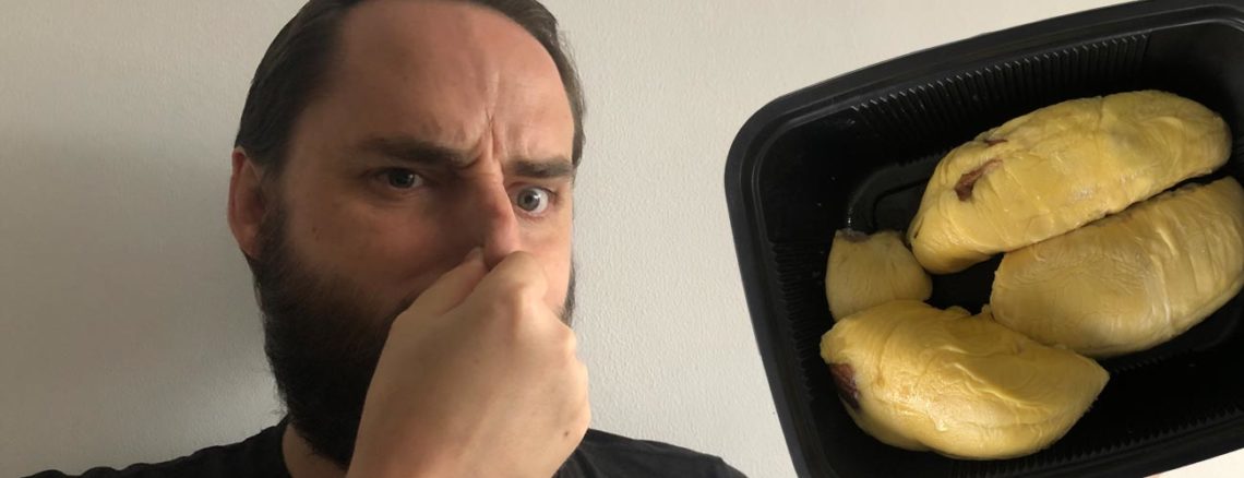 Image contains a photo of a bearded white man pinching his nose and looking unhappy. To the right of the man, there is a black plastic tray of durian, which is a yellowish fruit shaped in large pieces.