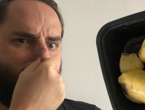 Image contains a photo of a bearded white man pinching his nose and looking unhappy. To the right of the man, there is a black plastic tray of durian, which is a yellowish fruit shaped in large pieces.