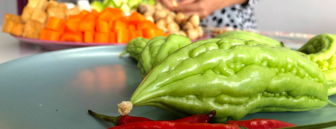 Image contains a photo of a number of vegetables. In the foreground, there are red chilis and green bitter melons of various sizes. In the background, there are cubed carrots, mushrooms, and more. A woman's hands can be seen near the vegetables in the background.