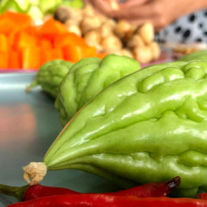 Image contains a photo of a number of vegetables. In the foreground, there are red chilis and green bitter melons of various sizes. In the background, there are cubed carrots, mushrooms, and more. A woman's hands can be seen near the vegetables in the background.