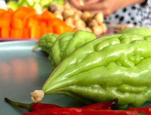 Image contains a photo of a number of vegetables. In the foreground, there are red chilis and green bitter melons of various sizes. In the background, there are cubed carrots, mushrooms, and more. A woman's hands can be seen near the vegetables in the background.