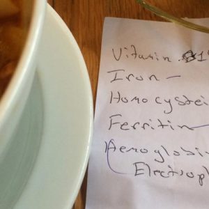 Image contains a photo of a white bowl of soup and noodles on the left, a glass cup of tea in the top right-hand corner, and a paper note in the bottom right-hand. On the note, there is handwriting that says "Vitamin B12, iron, homocysteine, ferritin, and hemoglobin electrophoresis."