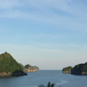 Image contains a photo of a harbour with a number of small hilly islands covered in jungle in the background. Above the islands, there is a blue sky with thin wisps of clouds.