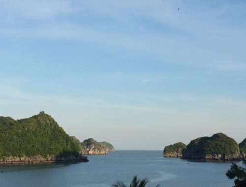 Image contains a photo of a harbour with a number of small hilly islands covered in jungle in the background. Above the islands, there is a blue sky with thin wisps of clouds.