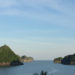 Image contains a photo of a harbour with a number of small hilly islands covered in jungle in the background. Above the islands, there is a blue sky with thin wisps of clouds.