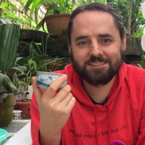 Image contains a photo of a bearded white man in a red hoodie sitting and holding a small tea cup. The man is smiling at the camera and there are plants surrounding him. To the left of he man, there is a white door being used as a table with a numbed of potted plants resting on it.